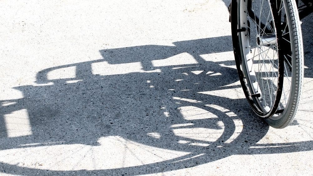 Side of a wheelchair, mainly the wheel, giving off a shadow of the wheelchair on the ground.