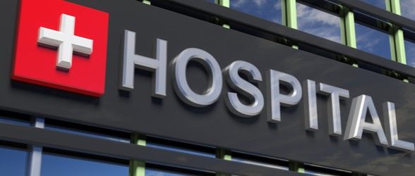 A close-up of a black sign on a building that says "Hospital" in gray letters. Next to the text is a red square with a gray cross symbol on it.
