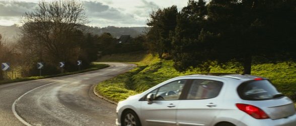 A silver car driving on an open road surrounded by trees, greenery, and a grayish sky.