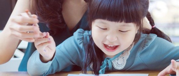 Young female-presenting, Asian-presenting child working on a drawing, with a female-presenting mother-figure sitting behind her, mostly not visible, guiding her hand.