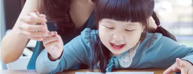 Young female-presenting, Asian-presenting child working on a drawing, with a female-presenting mother-figure sitting behind her, mostly not visible, guiding her hand.