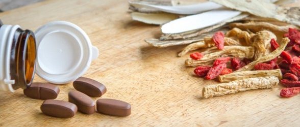A pill bottle with brown pills spilled out next to it. To the right of this is a pile of medicinal herbs.