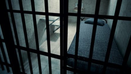 A view of a single-occupancy prison cell through the metal bars. Inside is a bed, toilet, and sink.