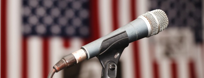 A photo of a microphone on a stand in front of blurred American flags in the background.