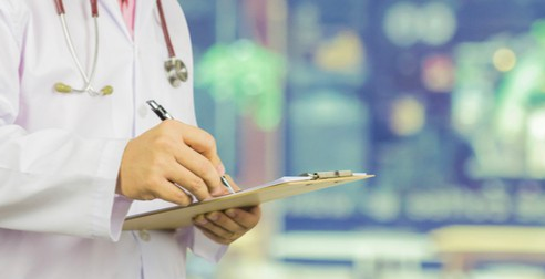 Torso of person in white lab coat with stethoscope around neck. The person is holding a clipboard and writing on it. The background is blurred.