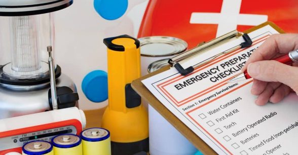 Emergency supplies including flashlight, radio, and waterbottle. A person is writing on an emergency preparedness checklist attached to a clipboard