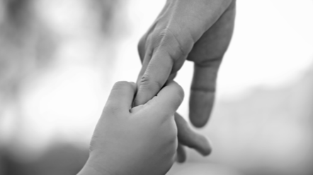 An adult hand holds a child's hand against a blurred background. Photo is black and white.