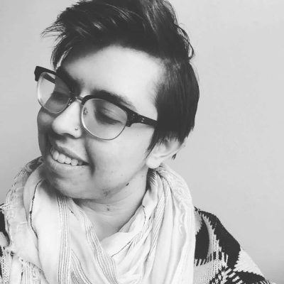 Sam Dylan Finch, a white femme and trans man, is shown here wrapped in a scarf and looking off to the side with his eyes nearly closed. He's smiling.