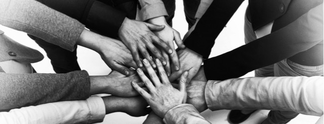A black and white photo of hands of different skin tones piled together in a circle showing solidarity.