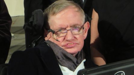 Photo of Stephen Hawking looking directly at camera. He is in a jacket and scarf and his assistive communication device is in front of him.