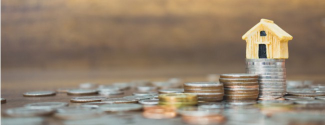Coins are scattered on a table. Three piles of coins, each bigger than the next, lead up to a stack of coins with a small wooden model of a house on top.