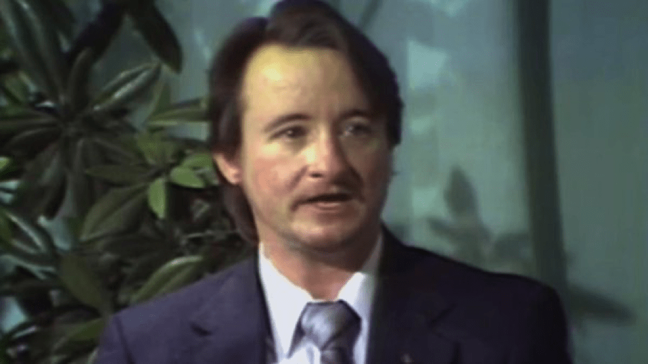 Lou Sullivan, a white trans man, sits in a TV studio. He is wearing a blue suit and tie.