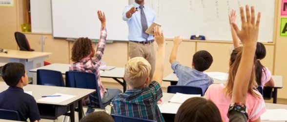 A teacher at the front of the class pointing who a student whose hand is raised. There is a group of students sitting in front of the teacher at their desks.