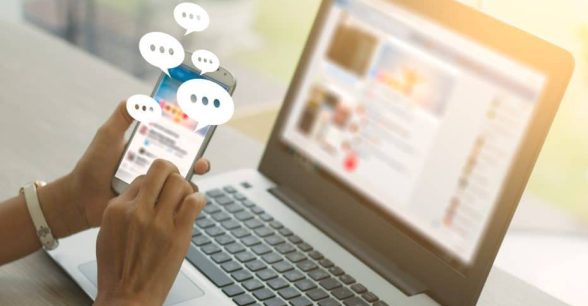 A person's hands holding a cell phone with message bubbles coming out of it, in front of a laptop.
