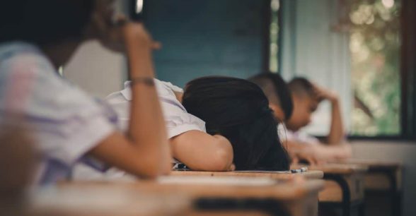 Four students sit at desks, one rests their head on their desk and the others rest their heads in their hands.
