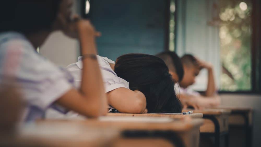 Four students sit at desks, one rests their head on their desk and the others rest their heads in their hands.