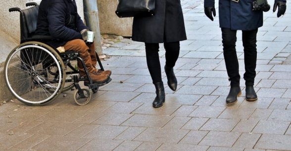 person sitting in a wheelchair holding cup for money next to people passing by