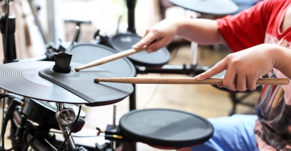 Photo of a young person playing drums. The person's face is not visible, but they are positioned behind the drum set.