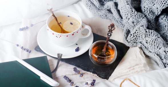 A cup of tea and pot of honey, a notebook and pen, and a blanket resting artfully on a bed, indicating relaxation.
