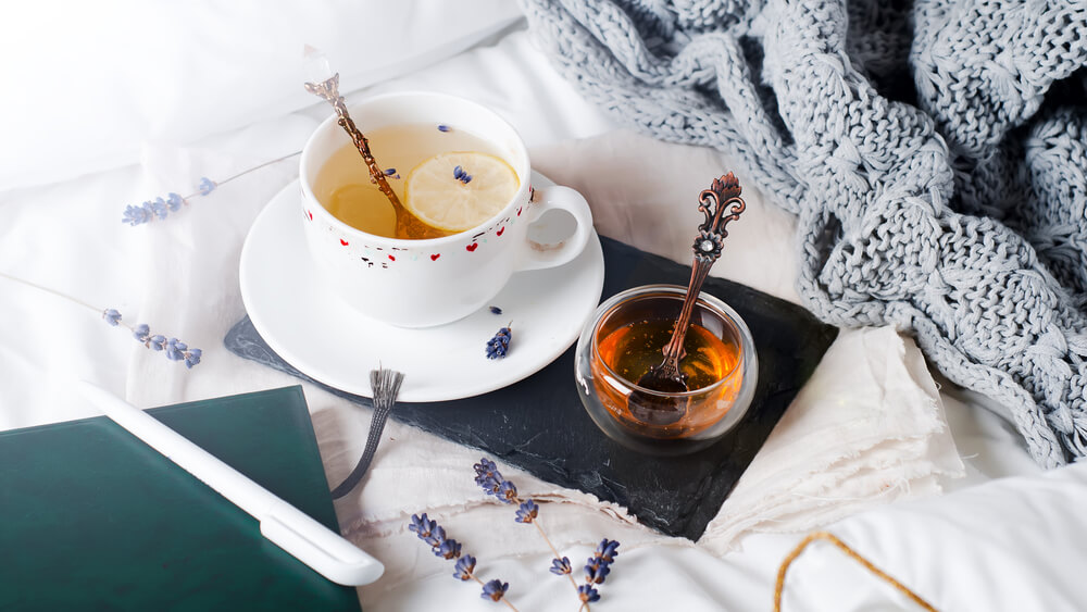 A cup of tea and pot of honey, a notebook and pen, and a blanket resting artfully on a bed, indicating relaxation.