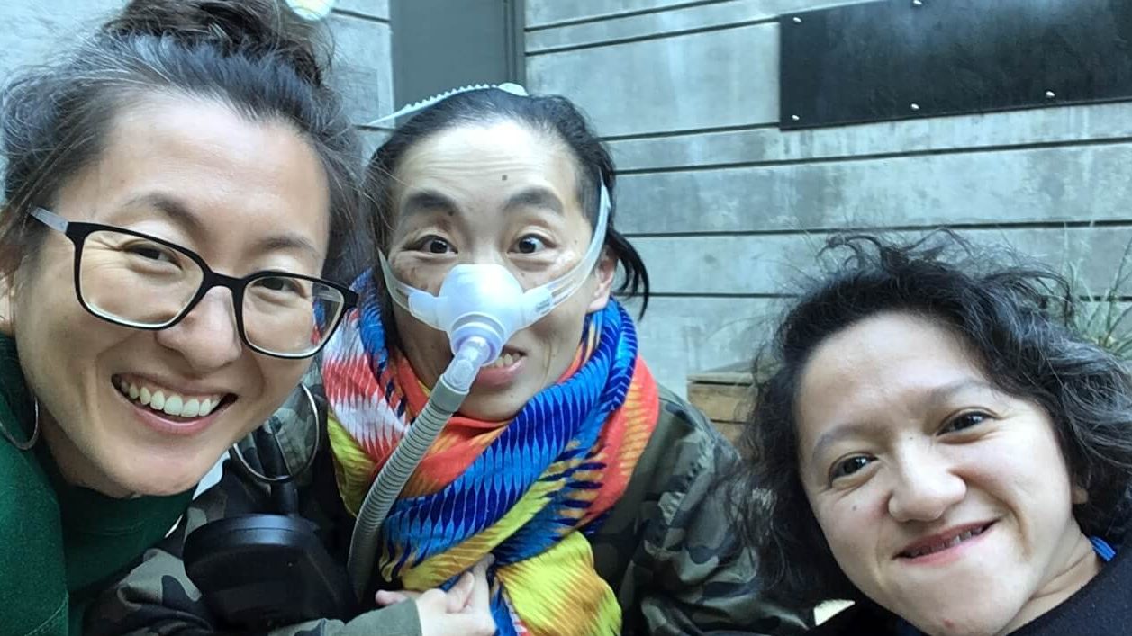 Photo of 3 disabled Asian American women, Mia Mingus, Alice Wong and Sandy Ho (from left to right). Mia is wearing glasses and large hoop earrings. Alice is wearing a brightly colored scarf and an army-camouflage-print jacket. She is wearing a mask over her nose with a tube for her Bi-Pap machine. Sandy has wavy short hair and is wearing a black sweater. Behind them is a concrete wall with a door.