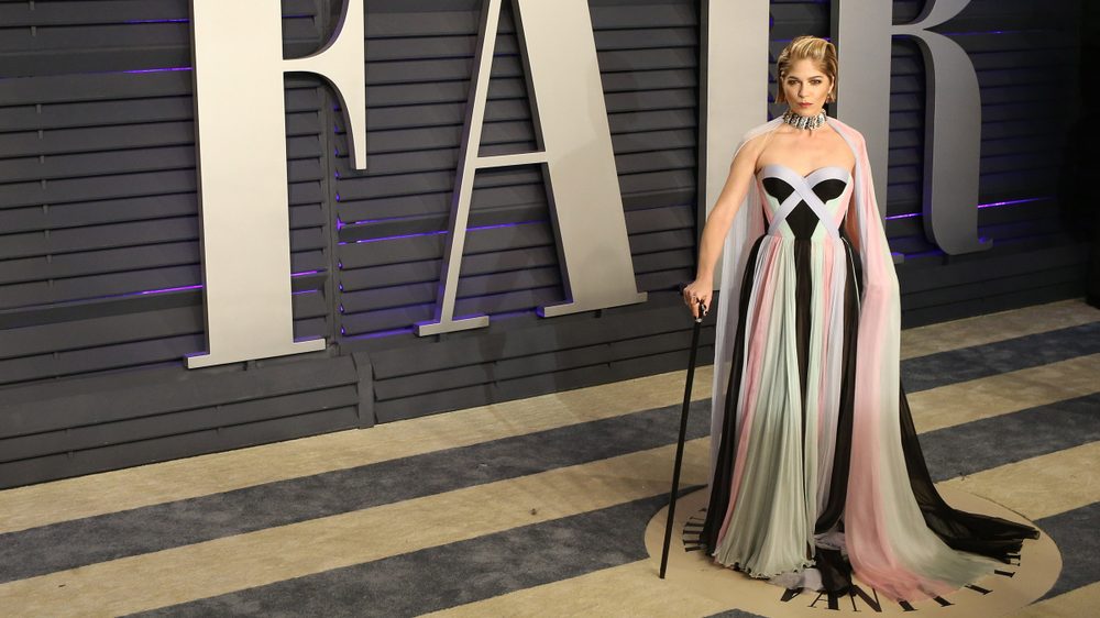 Selma Blair at the 2019 Oscars. She is glamorously dressed in a white, pink & white striped gown and holds a black cane.