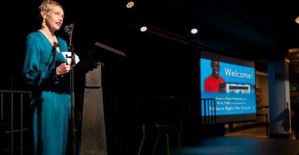 Photo of a woman in a blue jumpsuit standing at a podium on stage, speaking. She is looking up at the lights.