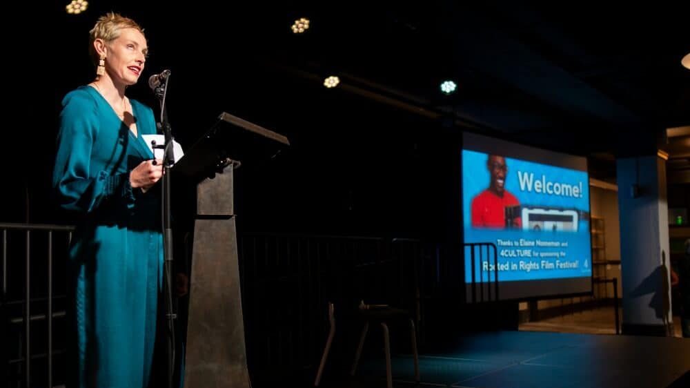 Photo of a woman in a blue jumpsuit standing at a podium on stage, speaking. She is looking up at the lights.