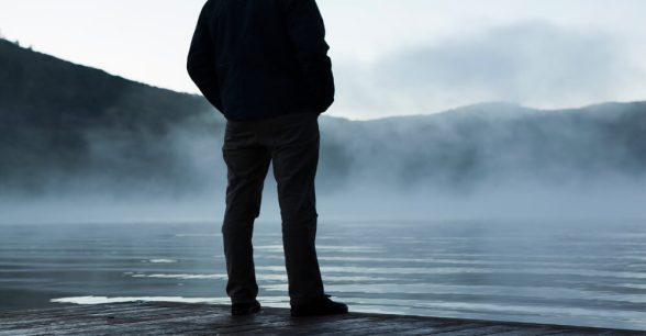 A person standing alone, looking out onto a hazy lake.