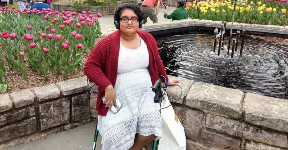 Noor, a south Asian transmasculine person in a white lace dress and red cardigan, sits on a green walker in front of a fountain and a large area of tulips on a sunny day