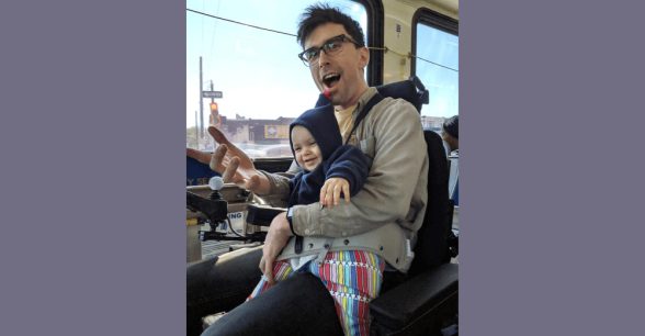 A photo of Liam sitting in his wheelchair on a bus, holding his daughter in his lap.