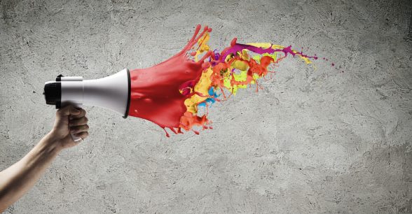 Hand holding a megaphone. The megaphone is white and red, and the red part fades off into multicolored paint splatters