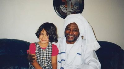 The author as a young child sitting on the lap of her Sudanese grandfather, who is dressed in a traditional white jellabiya and headwrap.