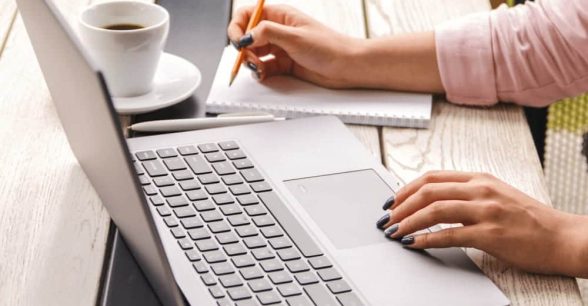 Closeup hands of white person in pastel pink shirt with painted nails working on laptop and making notes in their notebook, which is on wooden table with a laptop and a cup of coffee.