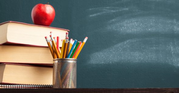 A pile of books with an apple on top and a cup with multi-colored pencils in it, in front of a chalkboard.