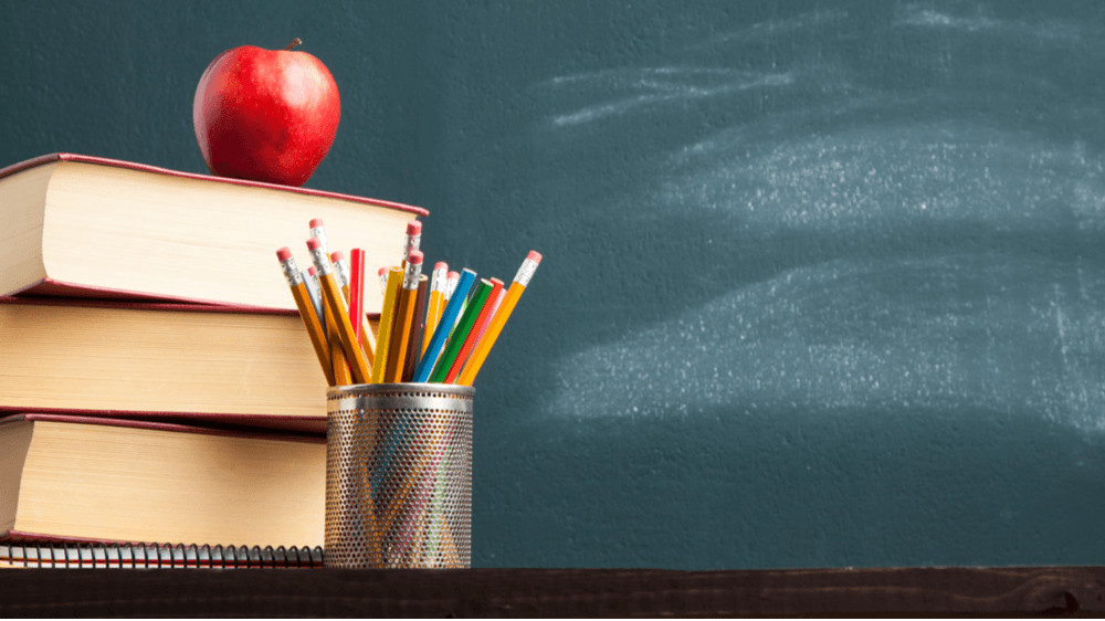 A pile of books with an apple on top and a cup with multi-colored pencils in it, in front of a chalkboard.