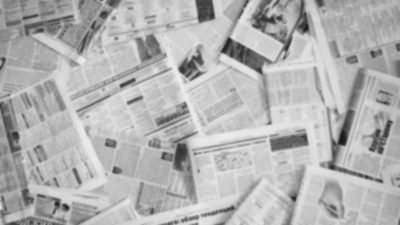 A blurred photo of a scattered pile of newspapers, printed all in black and white.