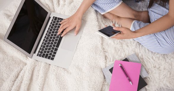 Top-down partial view of a white person sitting cross-legged on a bed, holding a cell phone, and resting her hand on their keyboard. There is a pink notebook and pen next to them.