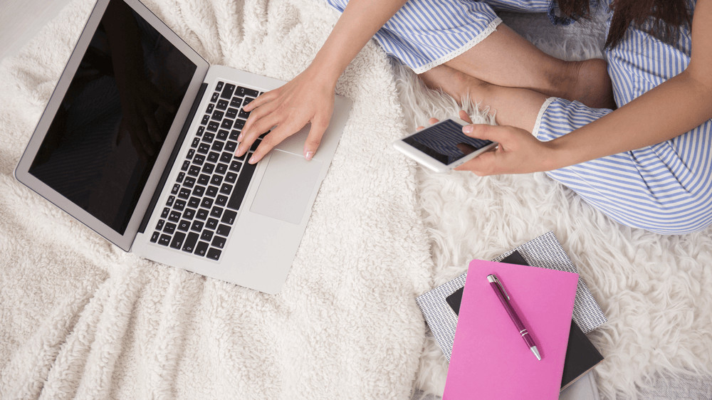 Top-down partial view of a white person sitting cross-legged on a bed, holding a cell phone, and resting her hand on their keyboard. There is a pink notebook and pen next to them.