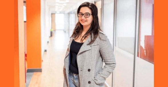 Malvika Padin standing in a well-lit hallway wearing black-rimmed glasses, shoulder-length dark brown hair left loose, clad in a black top, jeans and a grey coat with her hands in her coat pockets.