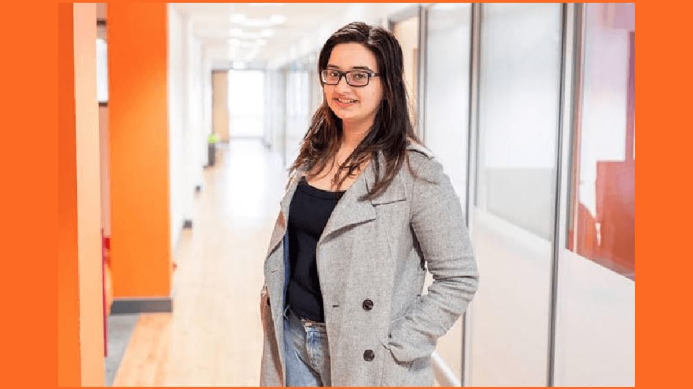 Malvika Padin standing in a well-lit hallway wearing black-rimmed glasses, shoulder-length dark brown hair left loose, clad in a black top, jeans and a grey coat with her hands in her coat pockets.