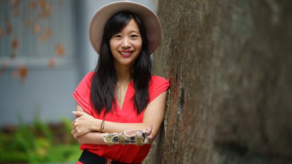Photo of Tiffany, an Asian woman with dark hair below her shoulders, leaning against a tree. She is wearing a hat and a red top. Her arms are crossed, and she is wearing a splint.