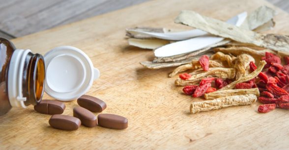 A pill bottle with brown pills spilled out next to it. To the right of this is a pile of medicinal herbs.