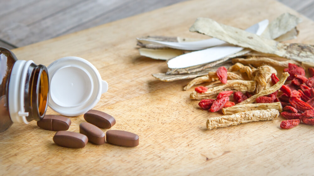 A pill bottle with brown pills spilled out next to it. To the right of this is a pile of medicinal herbs.