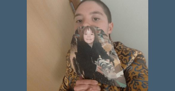 A young white woman with short hair, standing against a wall. In front of her face, she is holding a picture from when she was 8 years old, sitting in a wheelchair, smiling.