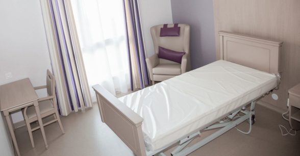 An empty room in a nursing home with a bed, chair, desk, and window.