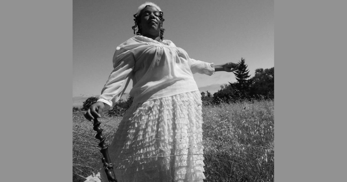 A grayscale photo of a Black woman in a flowy white top and skirt, holding a cane in one hand, her other hand outstretched. Her eyes are peacefully closed. She is standing in an overgrown field with trees behind her.