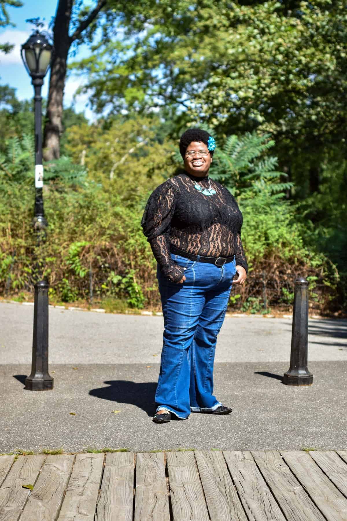 Denarii Grace stands outdoors on a sunny day, smiling at the camera in jeans and a black lace top, surrounded by trees..
