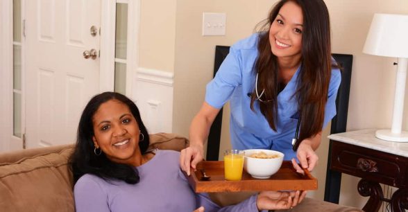 Two younger, dark-haired people with long hair - one brown-skinned, the other fairer skinned - in a living room. The fairer skinned person, wearing blue scrubs and a stethoscope around their neck, is standing beside the couch where the other person is seated. They're both smiling at the camera as the standing person passes a wooden tray with a white bowl of food and a glass of orange juice to the seated person.