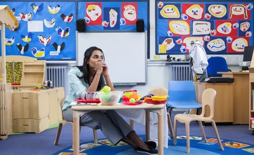 Stressed brown-skinned teacher with long, straight, dark hair in a colorful classroom, sitting at the toy table.